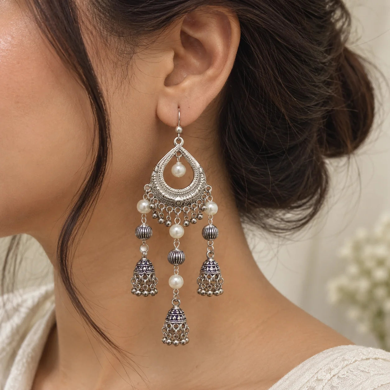 "Silver-toned statement
jhumka earrings with intricate teardrop motifs, white pearl accents, textured beads, and multiple cascading bell-shaped charms."