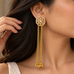 "Gold-toned floral earrings
featuring a round white centre stone, petal-shaped accents, a delicate pearl border, and multiple long cascading gold chain strands finished with small golden bead clusters."