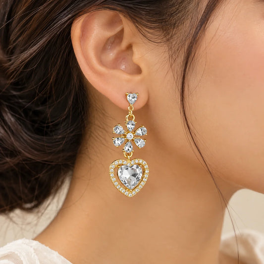 "Gold-toned dangle earrings
featuring sparkling floral crystal details and large heart-shaped clear crystal drops, displayed on a white jewellery card."
