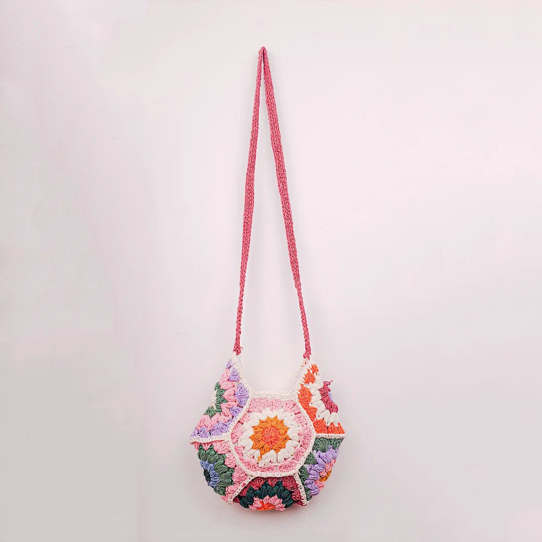 A colourful crochet bag made of hexagonal granny‑square patches in pink, orange, white, green, blue, and purple, with a mauve strap lying on other multicoloured crochet pieces.
