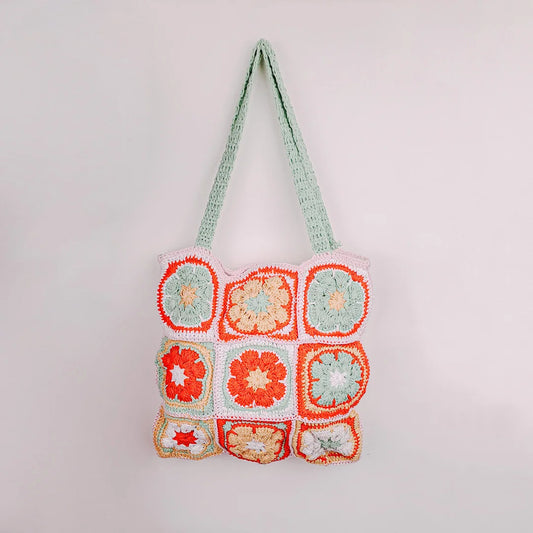 A crochet shoulder bag with nine granny‑square patches in red, green, and white floral patterns, lying on grass with a person’s knees in a white skirt visible in the top right corner.
