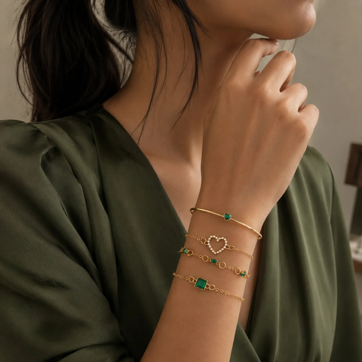"Gold-tone demifine bracelet
 stack featuring delicate chain designs with emerald-green accents, a heart-shaped detail, and a square-cut green stone centerpiece."
