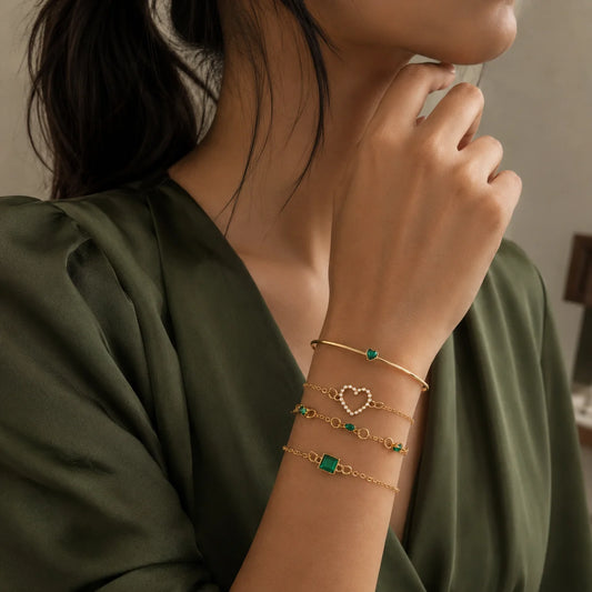 "Gold-tone demifine bracelet
 stack featuring delicate chain designs with emerald-green accents, a heart-shaped detail, and a square-cut green stone centerpiece."
