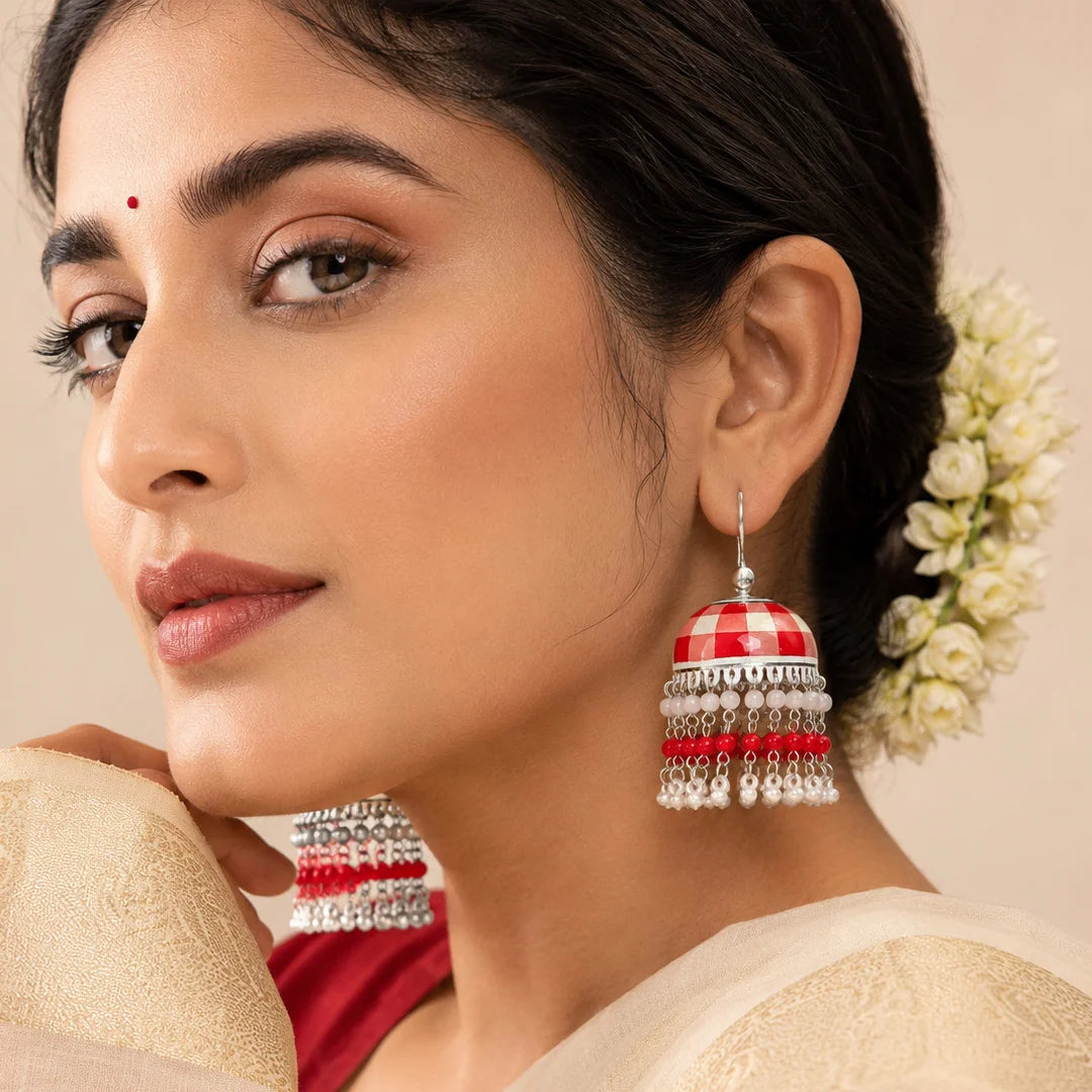 Red and white checkered dome jhumka earrings with silver ghungroo layers. Decorated with pearl and bead tassels in a cascading design. Traditional festive earrings displayed on a wooden background.

