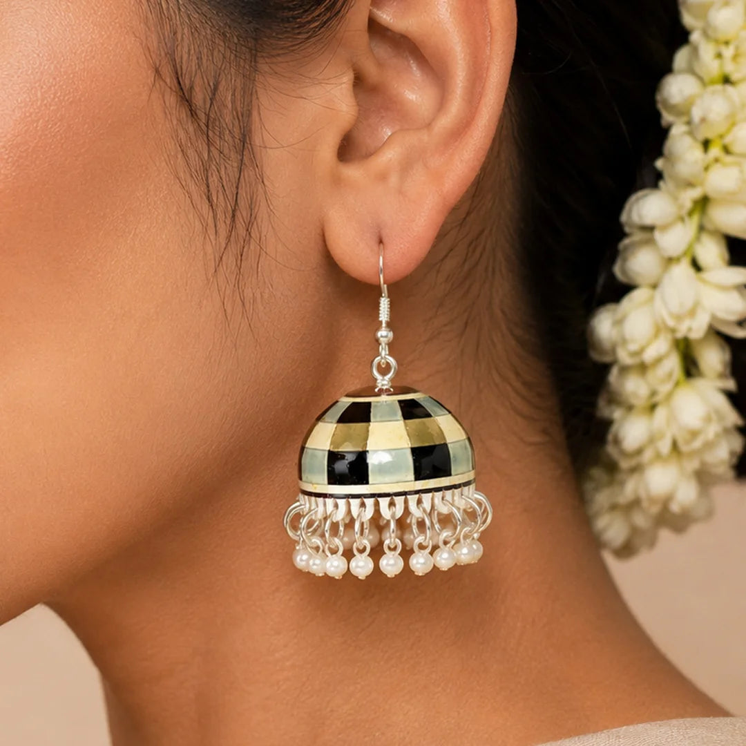 Pair of dome-shaped jhumka earrings with black, ivory, and grey checkerboard pattern and pearl bead fringe, placed on a wooden surface.
