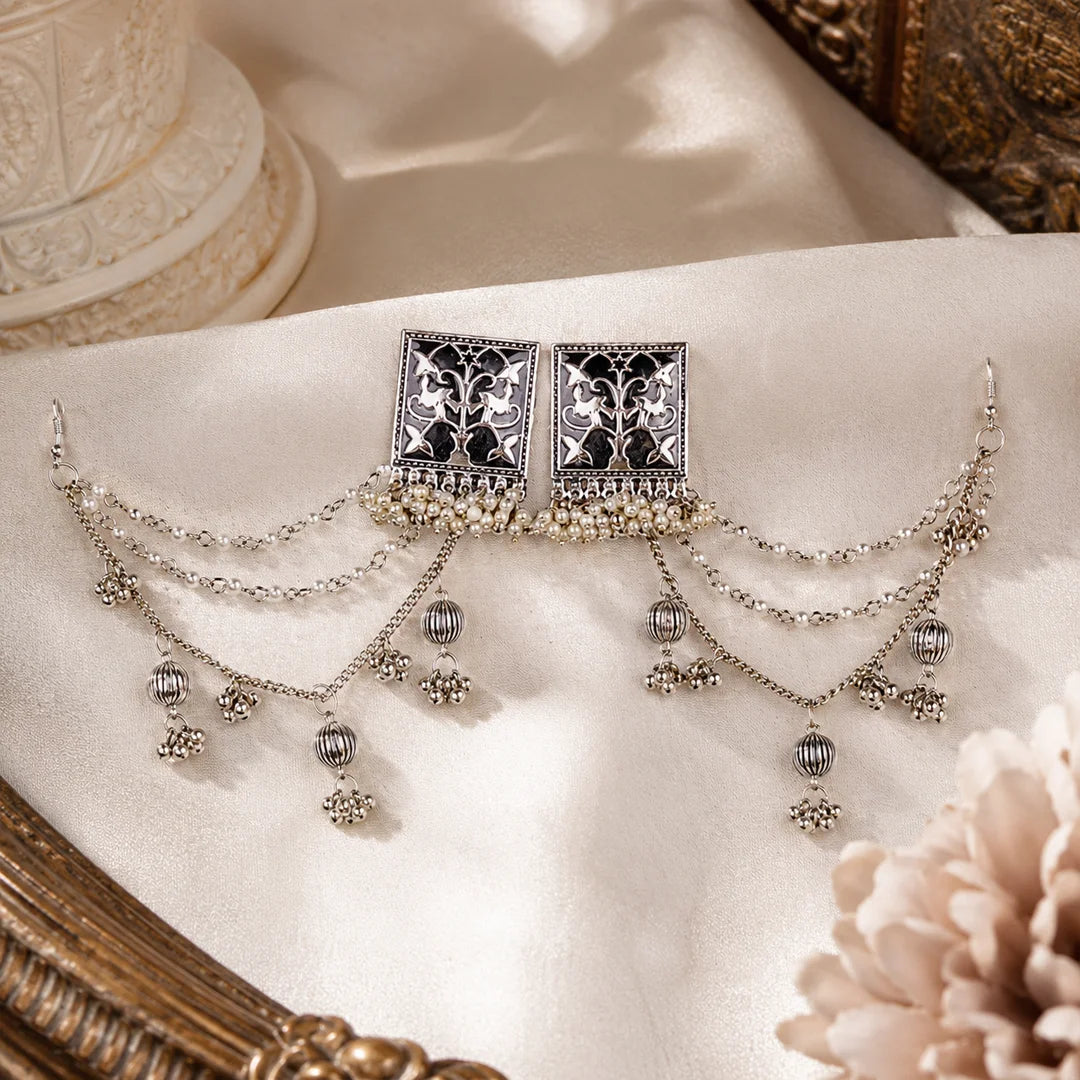 Oxidized silver earchain earrings with square floral studs, layered pearl chains, and jhumka-style danglers arranged on a wooden surface.
