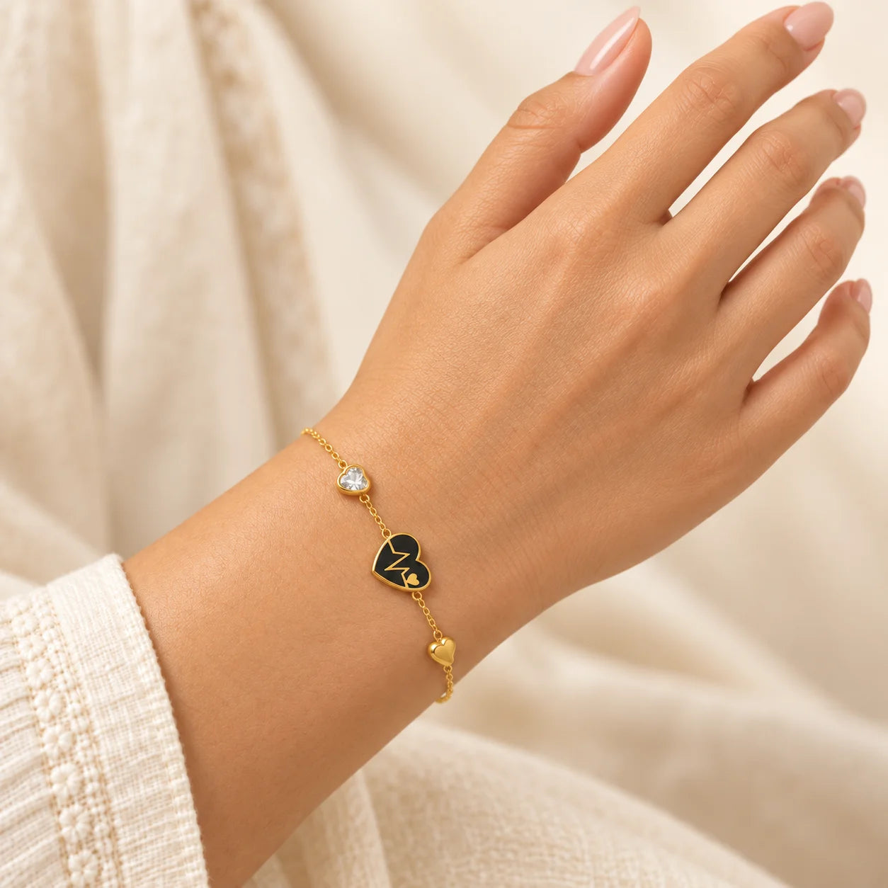 "Gold-tone chain bracelet
 featuring a small black heart-shaped charm, an adjustable chain extension, and a delicate heart-shaped end charm, worn on a wrist."
