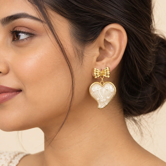 "Gold-tone bow
 earrings with glossy white heart-shaped drops featuring delicate raised detailing and pearl-like accents."
