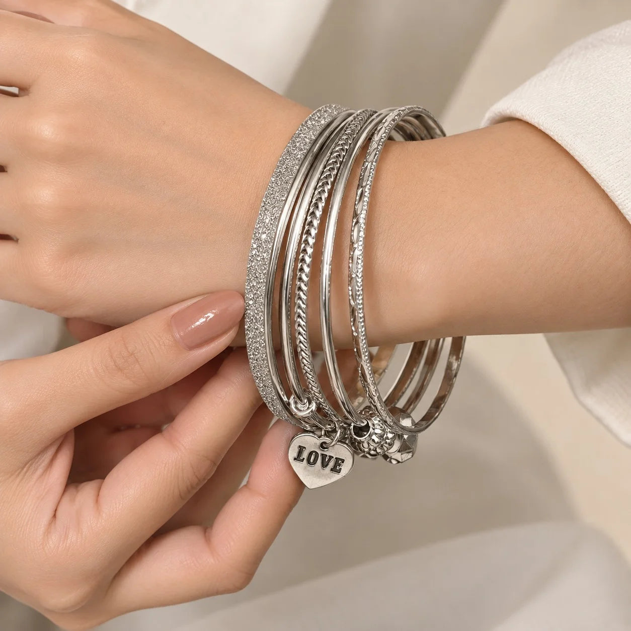 "Silver-tone bangle 
set featuring multiple textured and smooth bangles with a delicate heart-shaped charm, worn stacked around a wrist."
