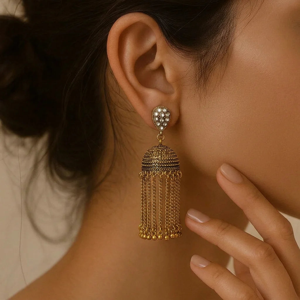 "Gold-tone 
 jhumka earrings featuring teardrop-shaped, rhinestone-embellished tops, textured dome-shaped drops, and long cascading golden chain tassels."

