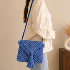 Powder blue handwoven macrame sling bag with open knot pattern and envelope flap. Features fringe trim, large knotted tassel, and braided rope strap with gold clasp. Boho cotton crossbody bag.
