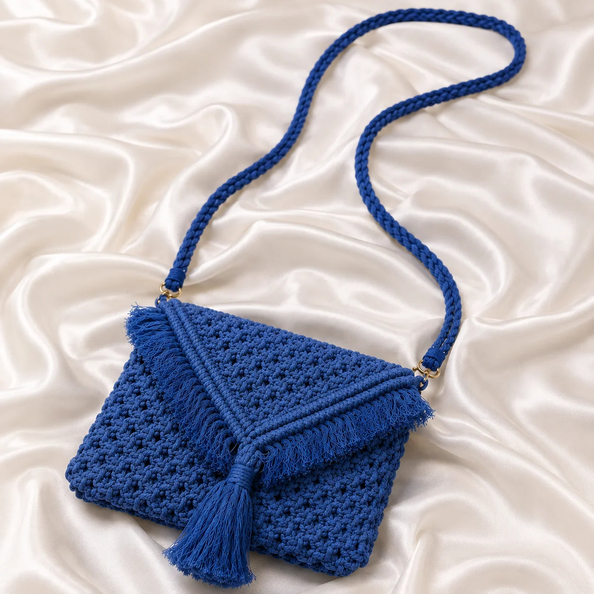 Royal blue handwoven macrame sling bag with open knot pattern and envelope flap. Features fringe trim, knotted tassel, and gold-tone strap ring. Boho cotton macrame crossbody bag.
