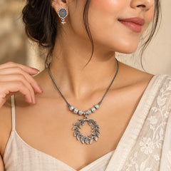 "Embrace nature-inspired elegance with this Silver Laurel Leaf Necklace Set, featuring a delicate silver-toned chain accented with textured beads and a beautifully sculpted circular leaf pendant. The open wreath-like design gives the pendant a graceful, organic feel, while the detailed leaf motifs add depth and character.

Paired with matching round stud earrings, this set offers a refined yet statement-making look. Style it with ethnic, fusion, or everyday outfits for an effortless touch of nature-inspired