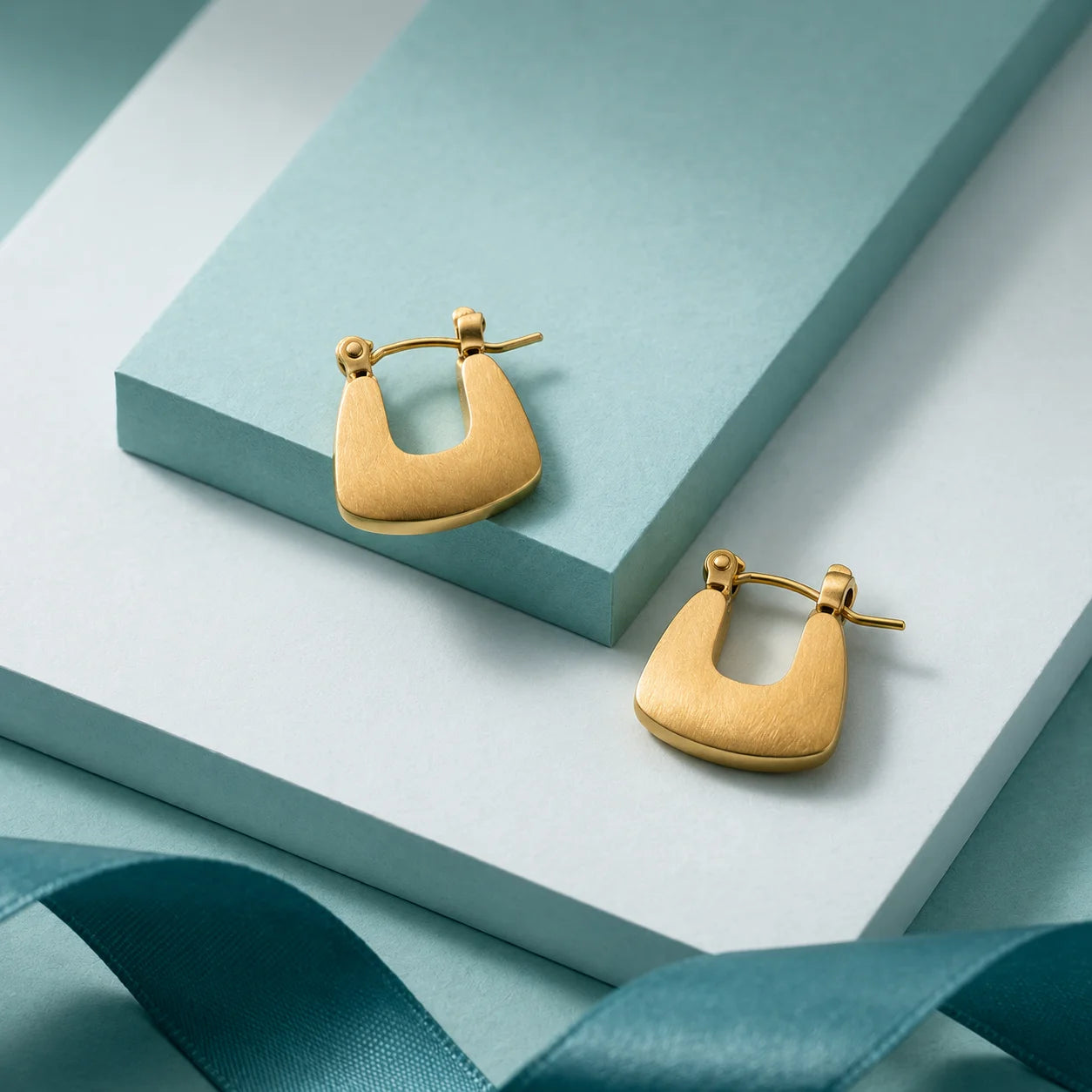 Pair of flat gold U-shaped huggie hoop earrings on wooden table. Minimalist gold-tone arch hoop earrings with hinged closure.
