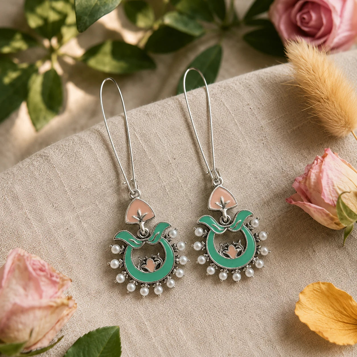 These traditional enamel hoop earrings feature hand-painted turquoise enamel work, delicate pink lotus detailing, and a halo of pearl beads on an silver chandbali frame. The wire hook design keeps them lightweight for all-day wear, while the vibrant enamel adds a festive pop to ethnic and indo-western outfits. Each piece reflects classic Indian craftsmanship with a modern boho twist.
