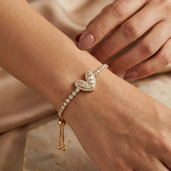 "Gold-tone bracelet 
featuring a delicate chain of sparkling clear crystals and an intricate heart-shaped pavé centerpiece, displayed on a wooden surface."
