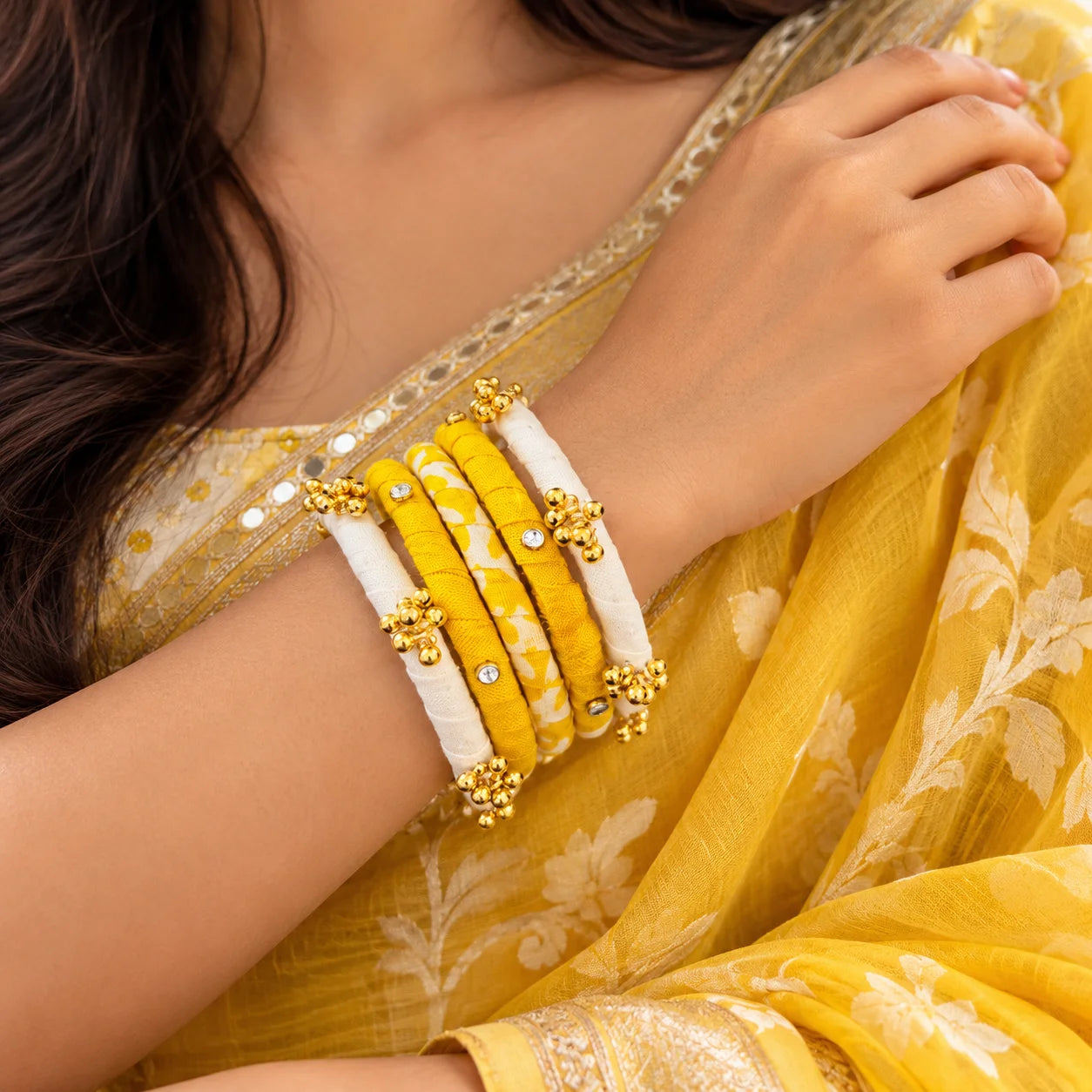 "Yellow and white 
fabric-wrapped bangle set featuring gold-tone bead cluster embellishments, decorative stone accents, and a mix of solid and printed designs, worn on the wrist."
