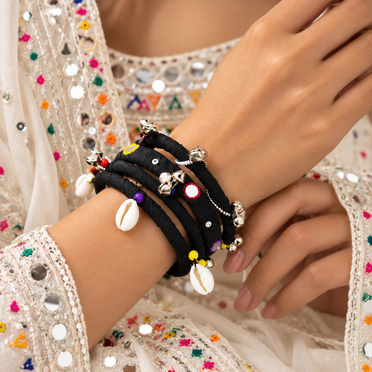 "Black fabric-wrapped
 bangle set featuring silver-tone bell charms, colourful beads, reflective mirror accents, and white shell embellishments, worn on the wrist."
