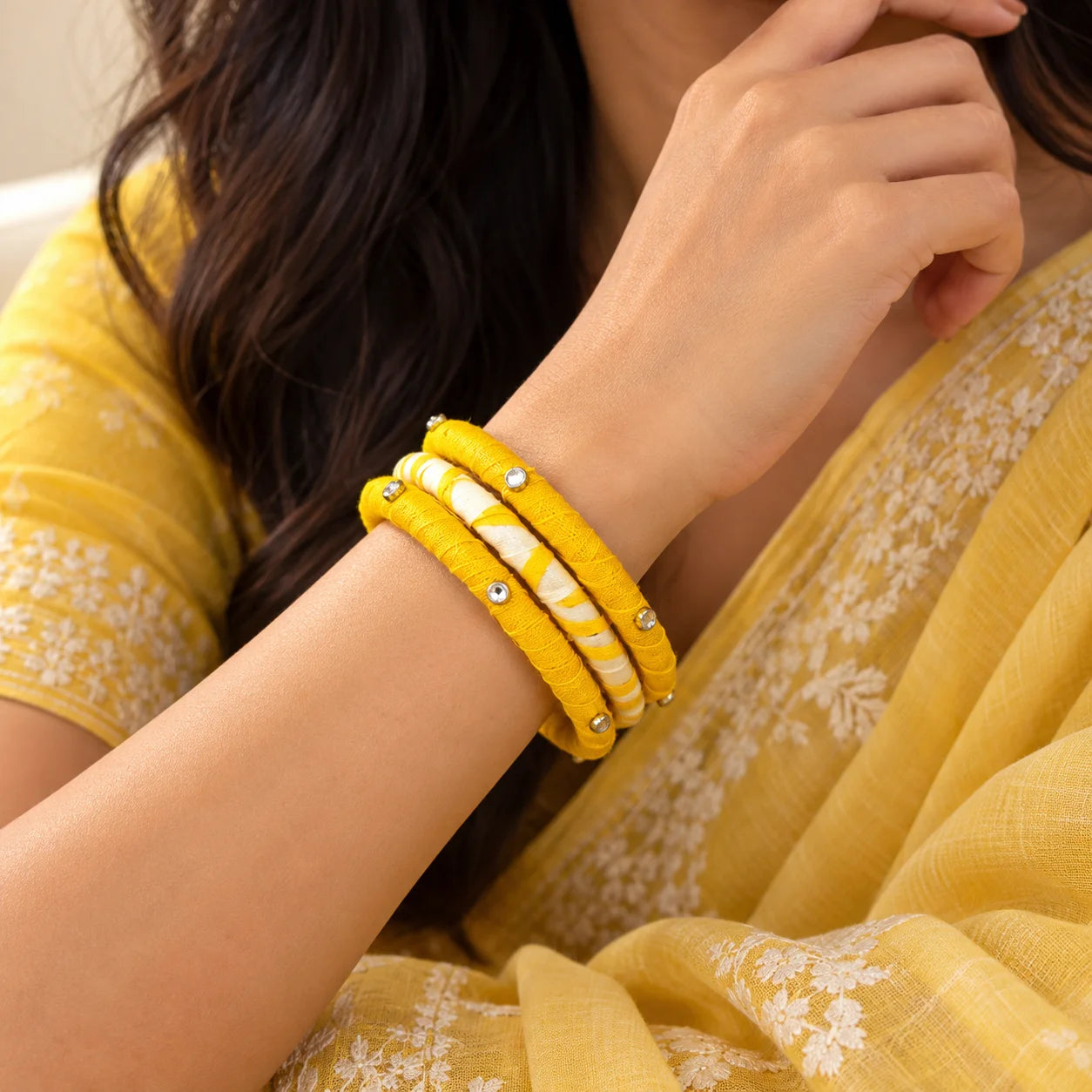 "Yellow and white fabric-wrapped 
bangle set featuring solid mustard yellow bangles, a white-and-yellow striped design, and silver-tone decorative stone embellishments."
