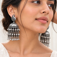 "Oxidised silver jhumka
 earrings featuring intricate peacock-inspired motifs, engraved dome-shaped drops, and multiple rows of dangling silver accents in a traditional ethnic design."
