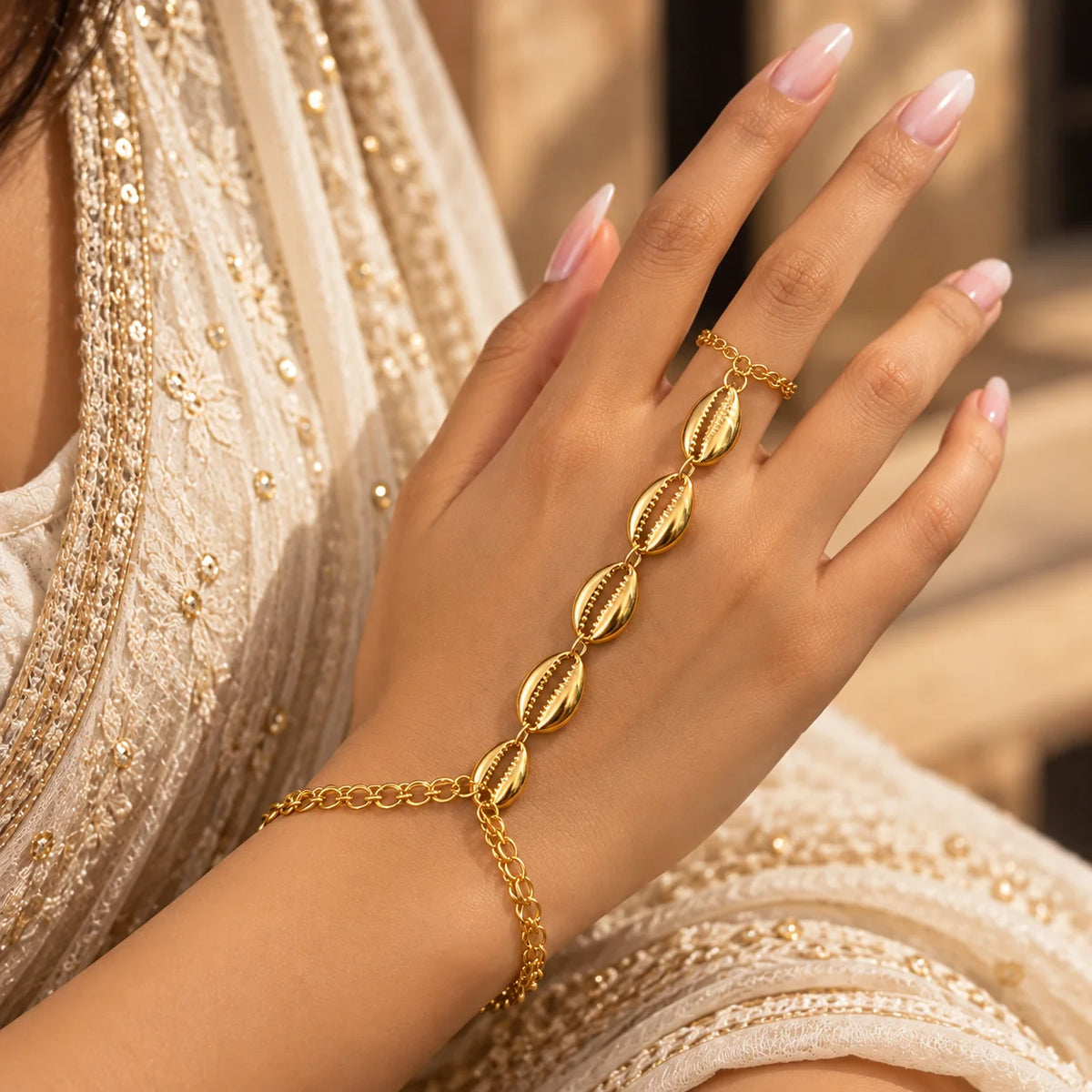 Gold-tone shell hand chain with four cowrie shell charms and delicate linked chains worn on the back of the hand.
