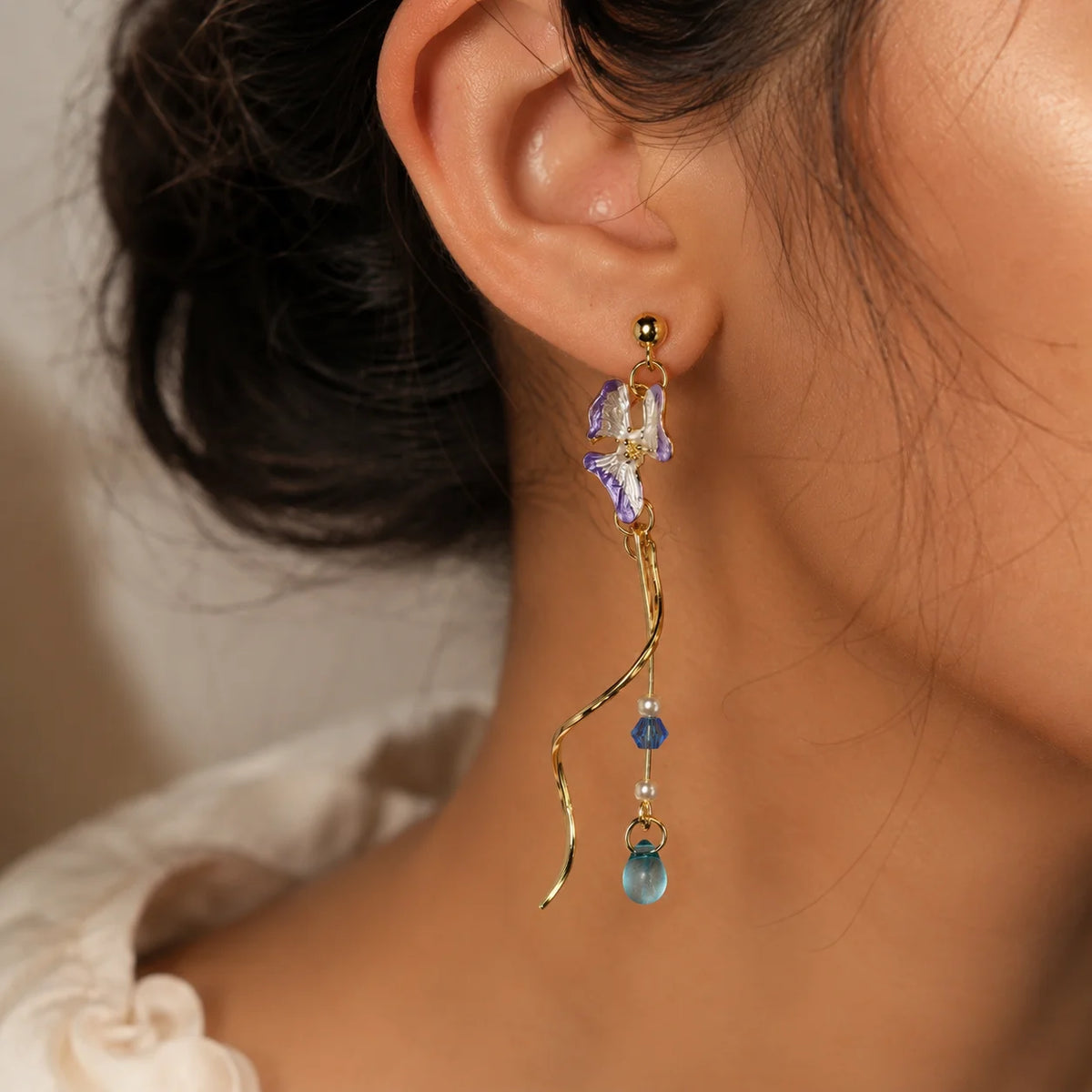 Pair of asymmetric gold-tone dangle earrings with lavender butterfly charms. Features blue crystal beads, faux pearls, and spiral bar details with teardrop ends. Mismatched western earrings with push-back posts on wooden surface.
