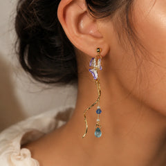 Pair of asymmetric gold-tone dangle earrings with lavender butterfly charms. Features blue crystal beads, faux pearls, and spiral bar details with teardrop ends. Mismatched western earrings with push-back posts on wooden surface.
