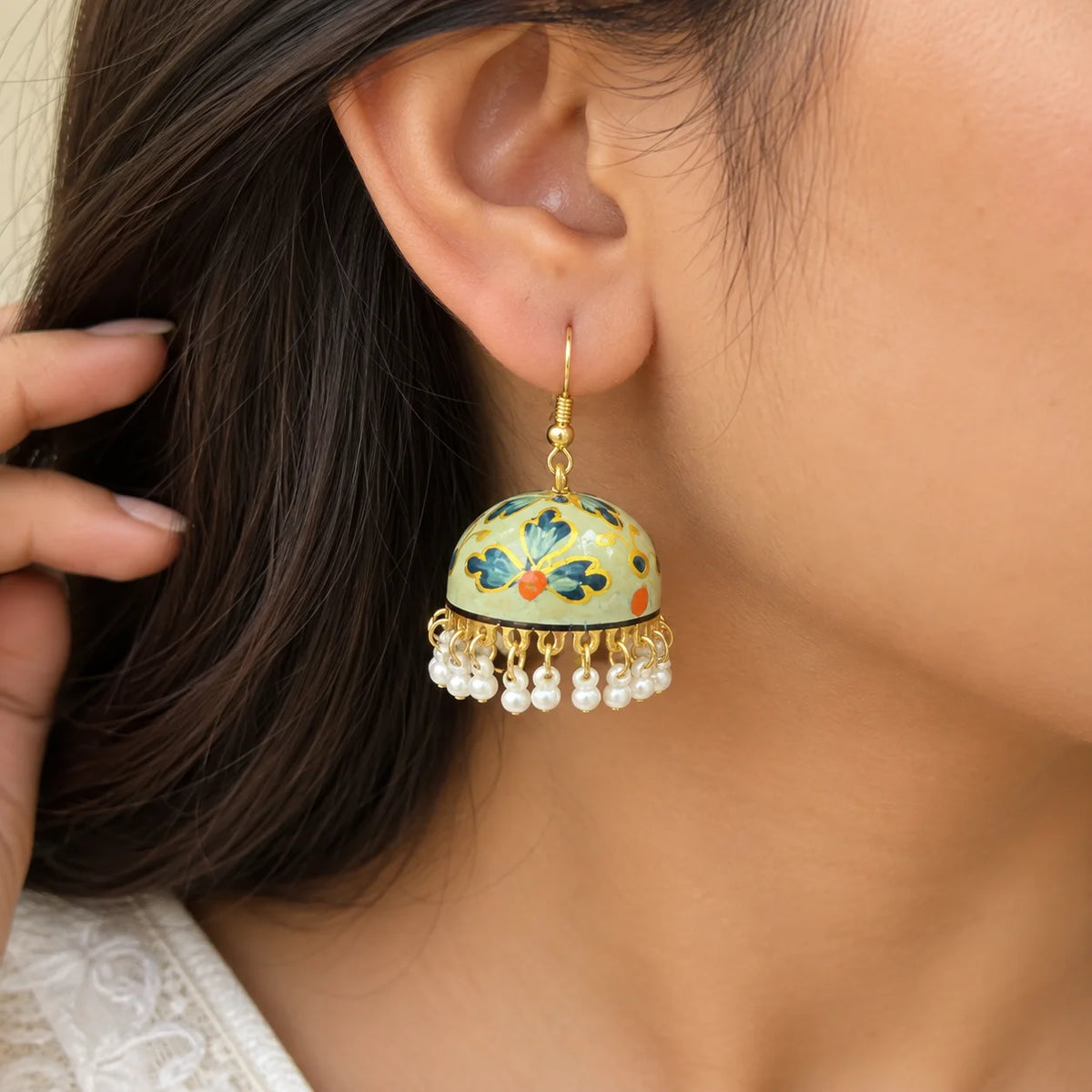"Sage-green dome-shaped
 jhumka earrings with blue floral and gold detailing, finished with white pearl-like bead danglers."
