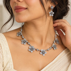 "Silver-toned floral necklace
 set featuring a delicate chain with intricate flower-shaped charms, paired with matching floral drop earrings on a clean background."
