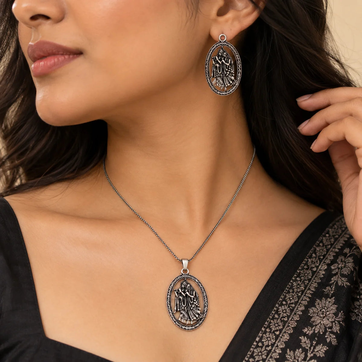 "Silver-toned Radha-Krishna
 necklace set featuring an intricately detailed oval pendant and matching oval earrings with ornate traditional borders."
