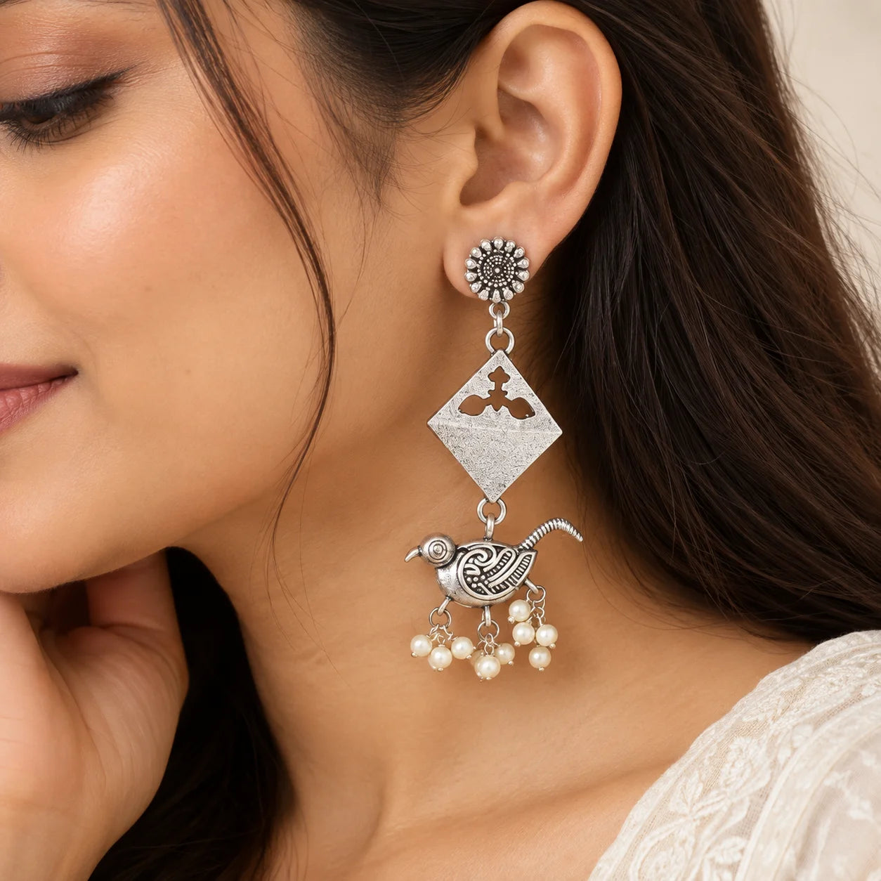 "Silver-toned statement 
earrings featuring intricate bird motifs, geometric diamond-shaped accents, and clusters of white pearl-like beads hanging at the bottom."
