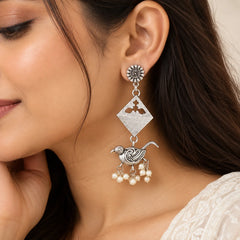 "Silver-toned statement 
earrings featuring intricate bird motifs, geometric diamond-shaped accents, and clusters of white pearl-like beads hanging at the bottom."

