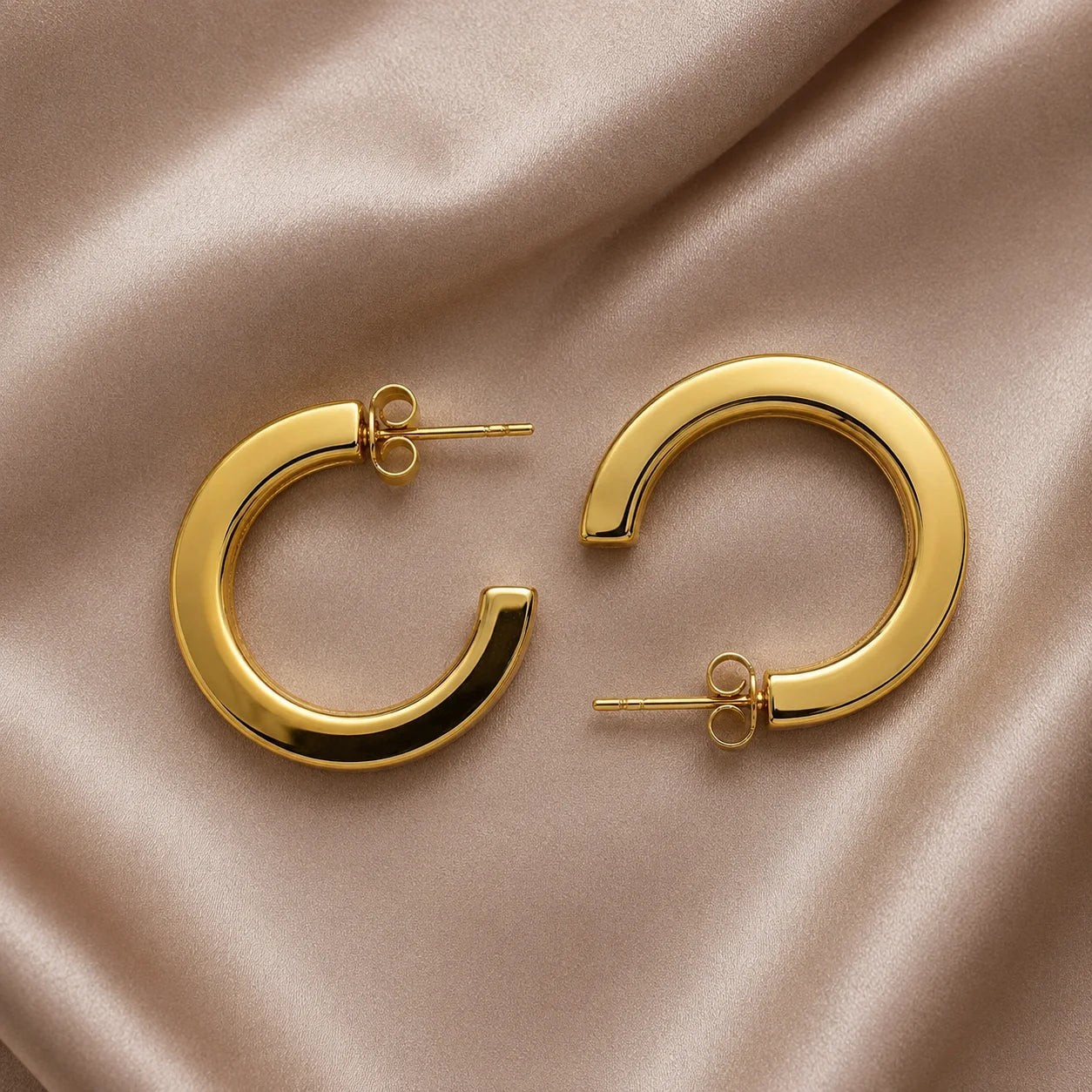 Pair of gold flat C-hoop earrings with push-back closure on wooden table. Chunky gold-tone open-ended huggie hoops with polished finish.
