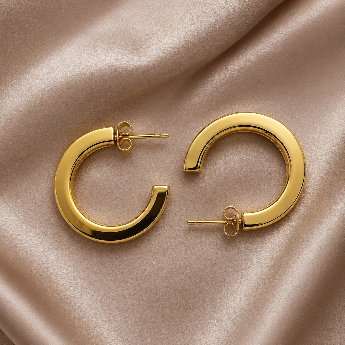 Pair of gold flat C-hoop earrings with push-back closure on wooden table. Chunky gold-tone open-ended huggie hoops with polished finish.
