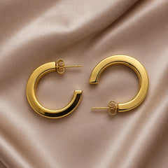 Pair of gold flat C-hoop earrings with push-back closure on wooden table. Chunky gold-tone open-ended huggie hoops with polished finish.
