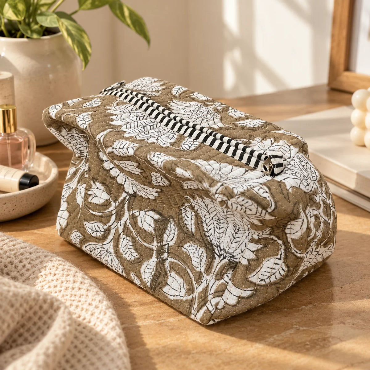 "Beige pouch with
 a white botanical floral print and contrasting black-and-white striped trim, photographed on a wooden table."
