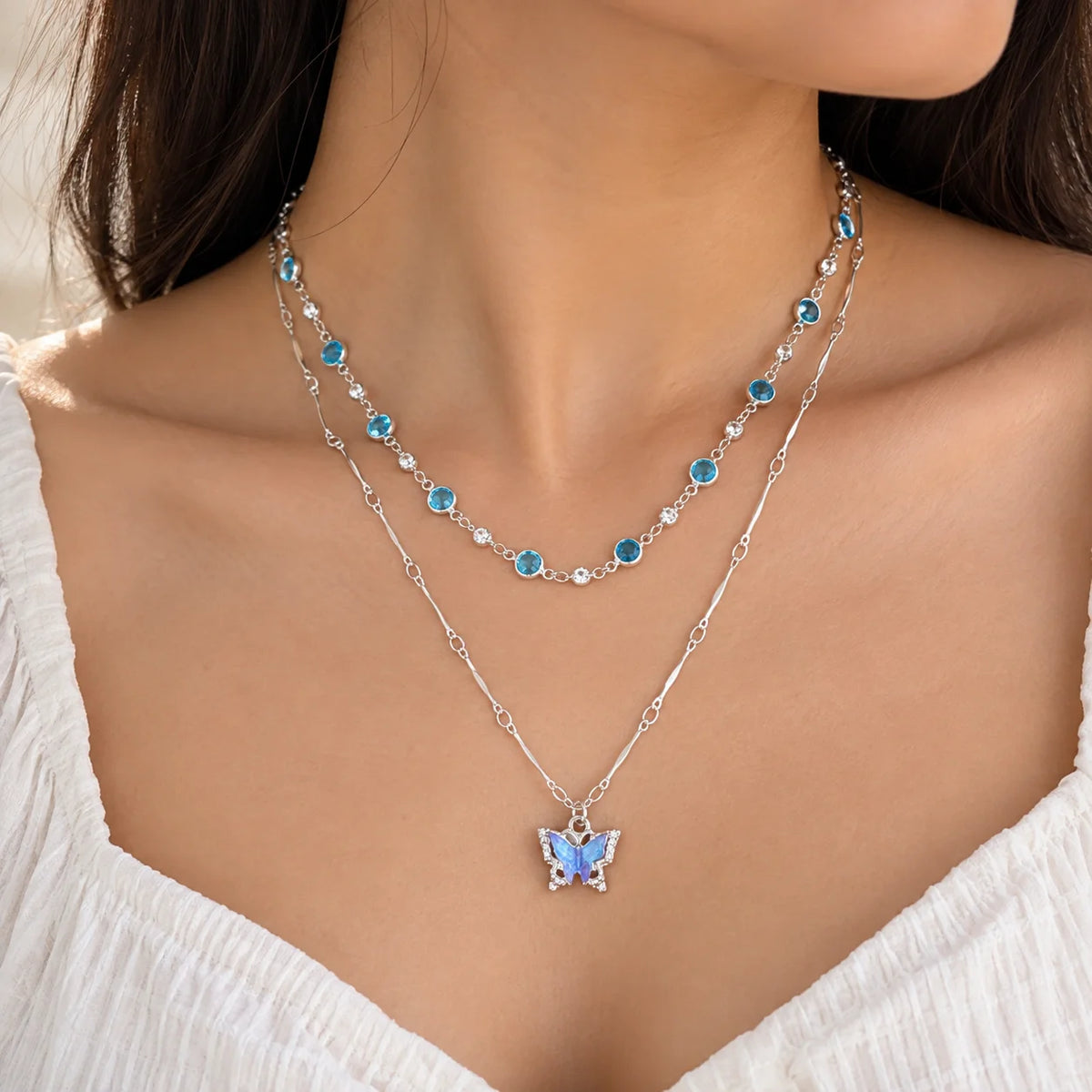 "Silver-toned layered 
necklace featuring aqua-blue crystal accents and a sparkling butterfly pendant, displayed on a dark background."
