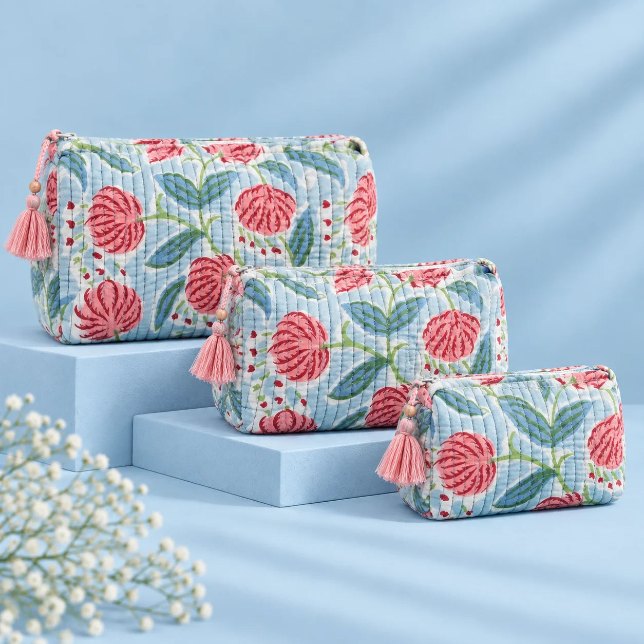"Set of three quilted
 fabric pouches with a white and grey base, featuring pink floral prints, green leaves, and decorative pink tassel accents."
