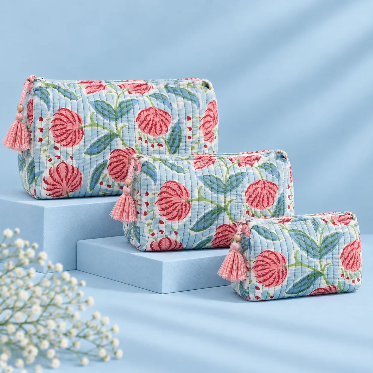 "Set of three quilted
 fabric pouches with a white and grey base, featuring pink floral prints, green leaves, and decorative pink tassel accents."
