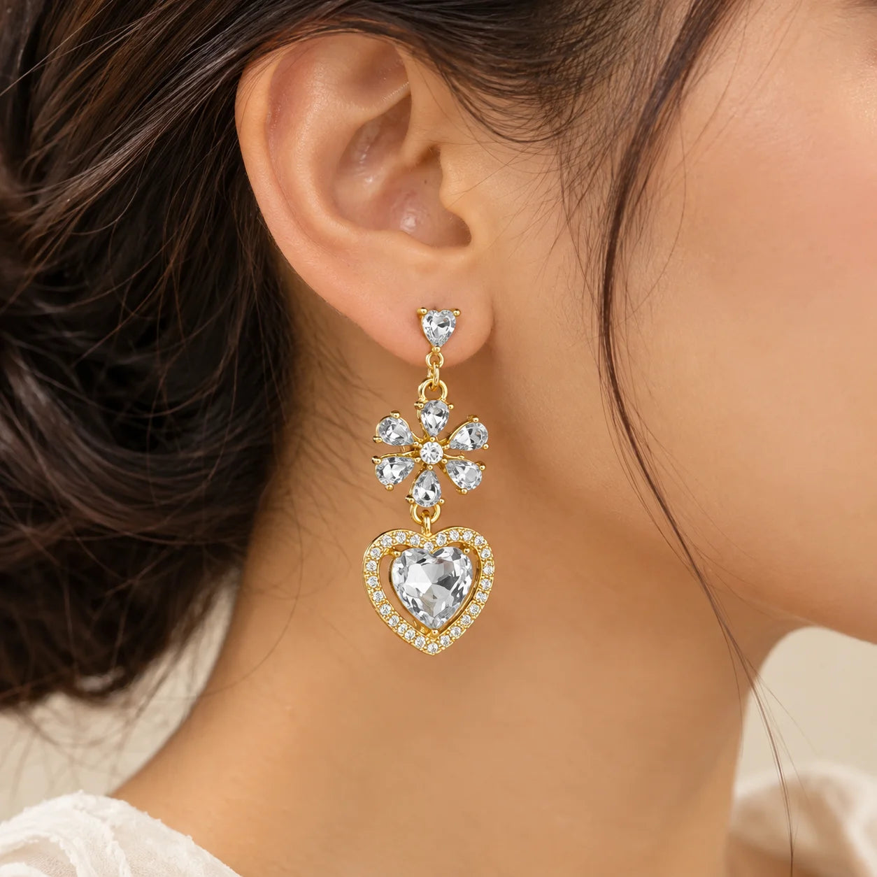 "Gold-toned dangle earrings
 featuring sparkling floral crystal details and large heart-shaped clear crystal drops, displayed on a white jewellery card."
