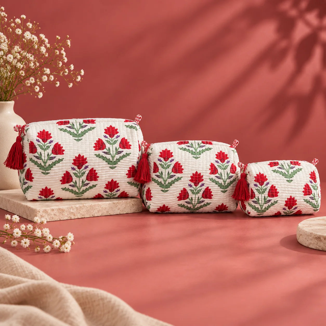 "Set of quilted fabric
 pouches with a white base, red floral and green leaf print, purple accents, and decorative red tassels."
