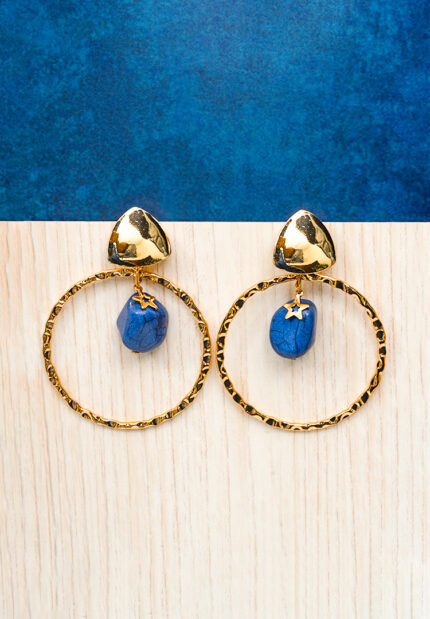 Chic metal hoop earrings featuring a polished gold-tone finish, dangling blue assymetrical stone, a golden charm and, a push back for easy wear.