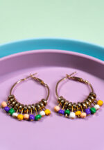 Chic metal hoop earrings featuring a polished gold-tone finish, multicolor finish featuring circular shaped golden beads, coloiurful dangling beads at the bottom and a hinged back.