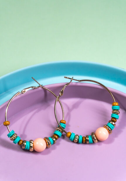 Chic metal hoop earrings featuring a polished gold-tone finish, playful turquoise accents, turquoise gold and wooden beads along with a soft pink coloured bead in the centre and a hinged back.