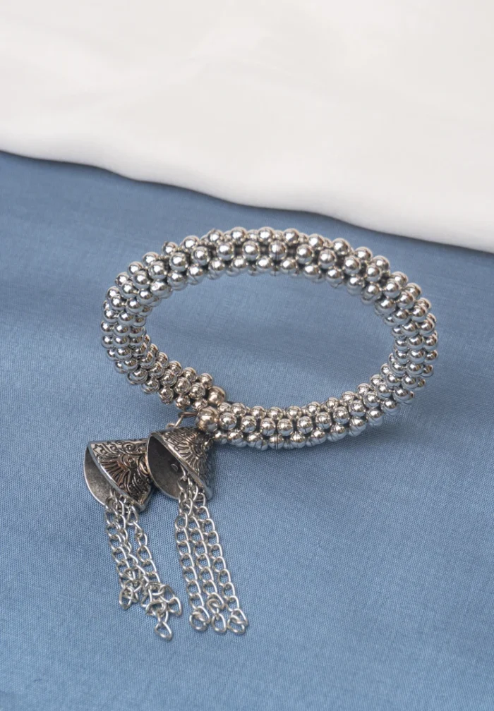 Beaded metal hinged bracelet in an oxidized silver finish, showcasing circular metallic beads and two ornate dangling bell-shaped charms with delicate chain tassels on each side.
