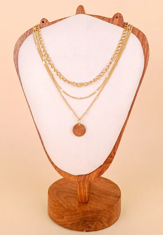 A pre-layered gold-toned necklace featuring three connected chains of varying lengths and textures, with a large, polished circular coin or medallion pendant hanging from the longest chain.