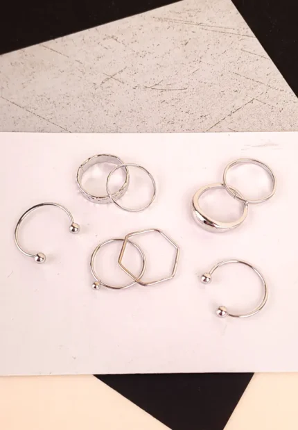 A collection of eight individual silver-toned stacking rings laid out on a black surface. The set includes two open cuff rings, a hexagon-shaped geometric ring, a thick band with crystal accents, and four thin polished bands of various sizes.