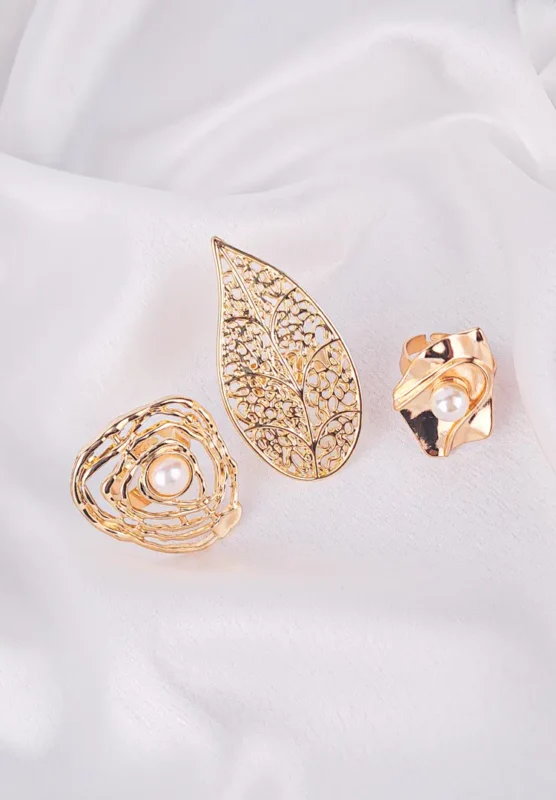 A trio of large, gold-toned statement rings. The set includes a wide etched leaf-shaped ring, a swirling abstract ring that cradles a pearl, and an asymmetrical eye-shaped ring also set with a pearl.