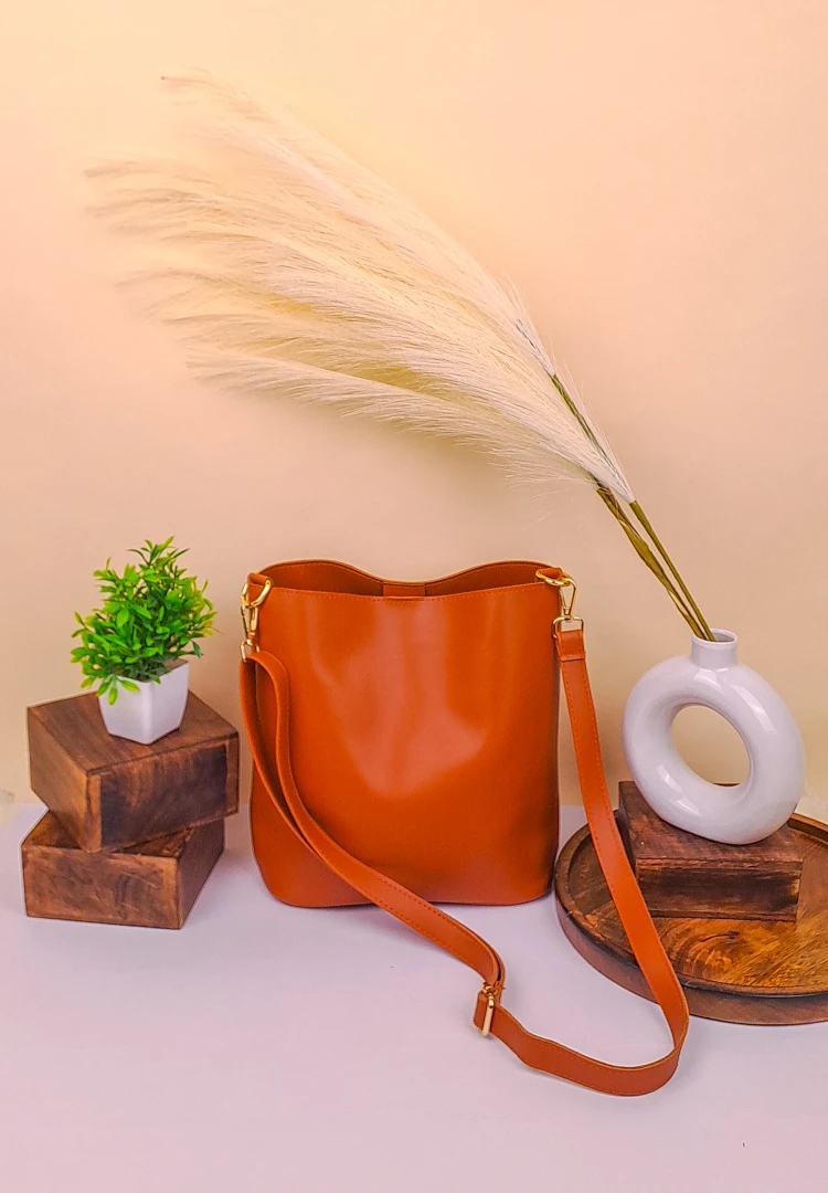 th (44) Tan bucket bag with wide shoulder strap, made of smooth faux leather, placed on a black table in a storage setting.