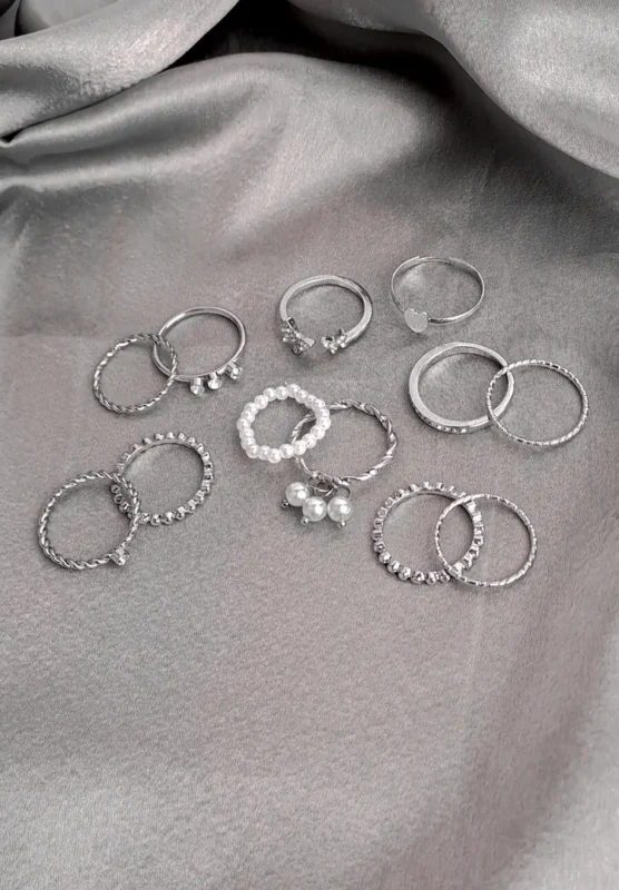 A large collection of twelve silver-toned stacking rings, including bands with pearl drops, continuous faux pearls, twisted rope texture, single crystal accents, and plain polished metal, scattered on a dark surface.